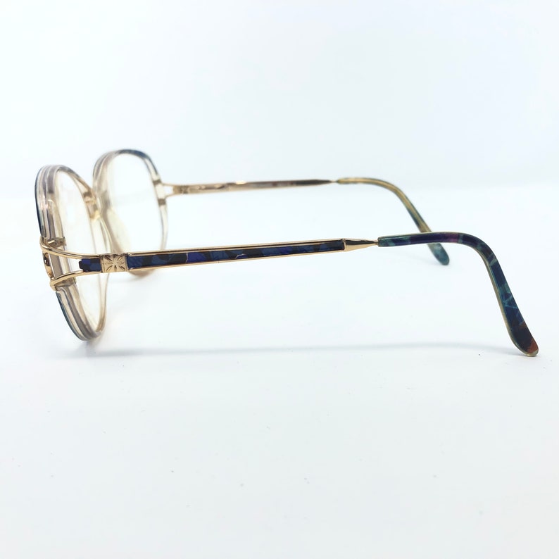 May include: Vintage eyeglasses with a gold-tone frame and clear lenses. The temples feature a dark blue and green marbled design. The glasses have a square shape and are set against a white background.