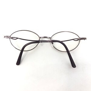 Peut inclure: Lunettes de forme ovale avec une fine monture en m&eacute;tal argent&eacute;. Les verres sont transparents et les branches sont marron fonc&eacute;. Les lunettes sont pos&eacute;es sur un fond blanc.