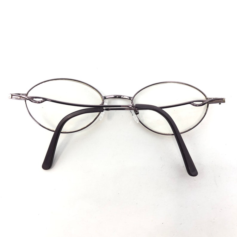 Peut inclure: Lunettes de forme ovale avec une fine monture en m&eacute;tal argent&eacute;. Les verres sont transparents et les branches sont marron fonc&eacute;. Les lunettes sont pos&eacute;es sur un fond blanc.