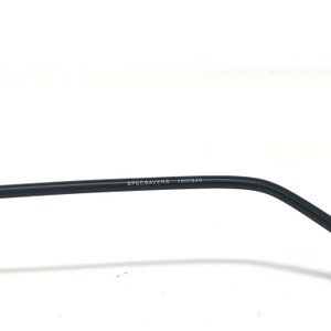 Puede incluir: Primer plano de una patilla de gafas negra. El texto "SPECSAVERS 1000840" est&aacute; impreso en blanco en la patilla. La montura est&aacute; sobre un fondo blanco.