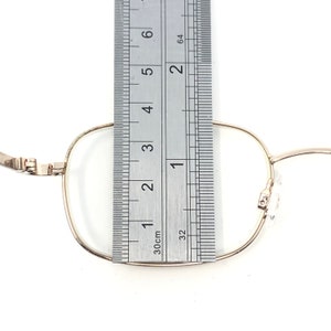 May include: Close-up of gold-rimmed eyeglasses with a ruler placed over the lens. The ruler shows measurements in centimetres. The glasses have a rectangular shape and are set against a white background.