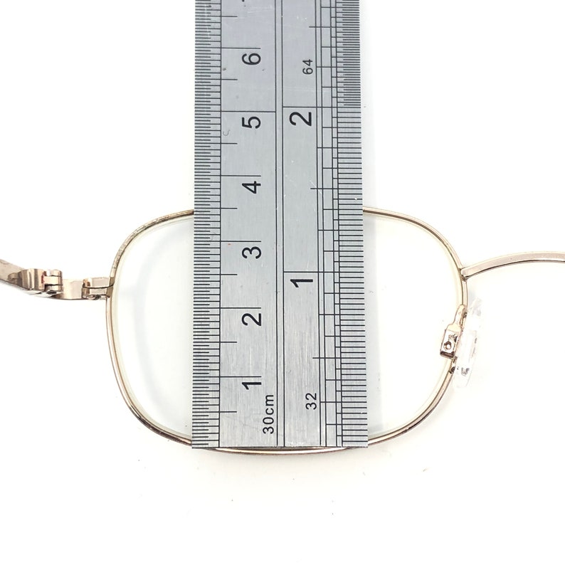 May include: Close-up of gold-rimmed eyeglasses with a ruler placed over the lens. The ruler shows measurements in centimetres. The glasses have a rectangular shape and are set against a white background.