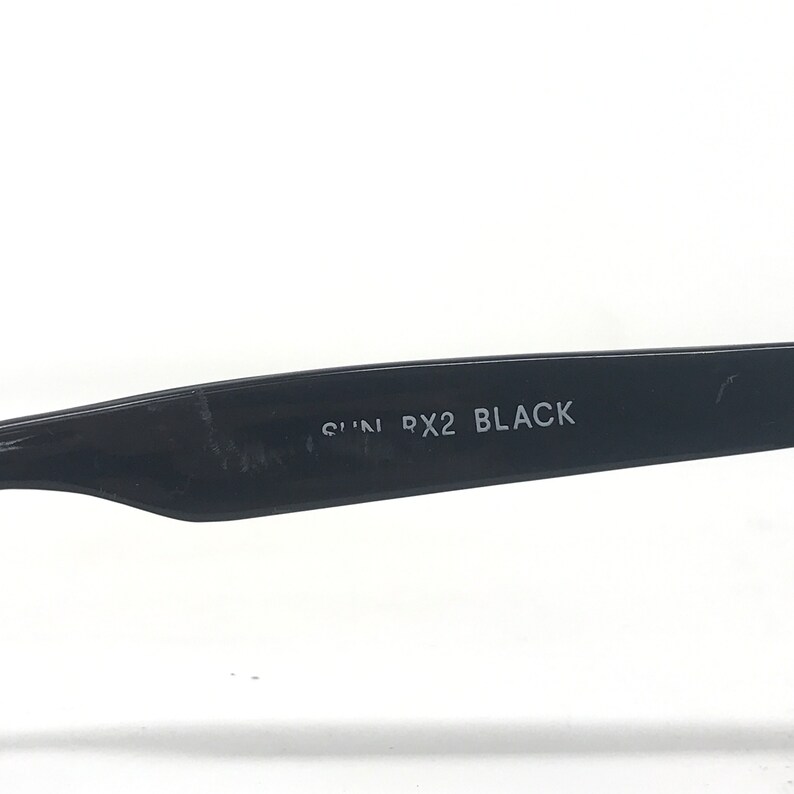 May include: Close-up of the arm of black sunglasses. The text "SUN PX2 BLACK" is printed in white on the black arm. The sunglasses are set against a white background.