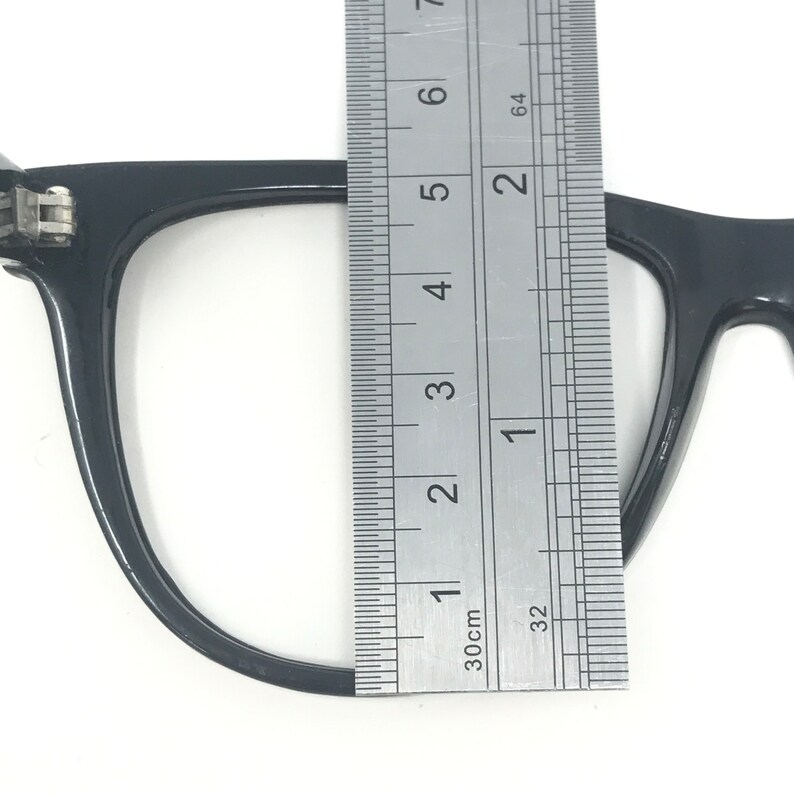 May include: Close-up of a black eyeglasses frame with a ruler measuring its height. The frame is approximately 2 inches tall. The eyeglasses are set against a white background.