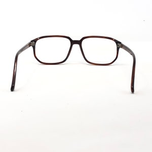 May include: Brown rectangular eyeglasses with clear lenses. The glasses have a classic, retro design with a dark brown frame and temples. The arms of the glasses are extended, showcasing the full shape and style of the eyewear.