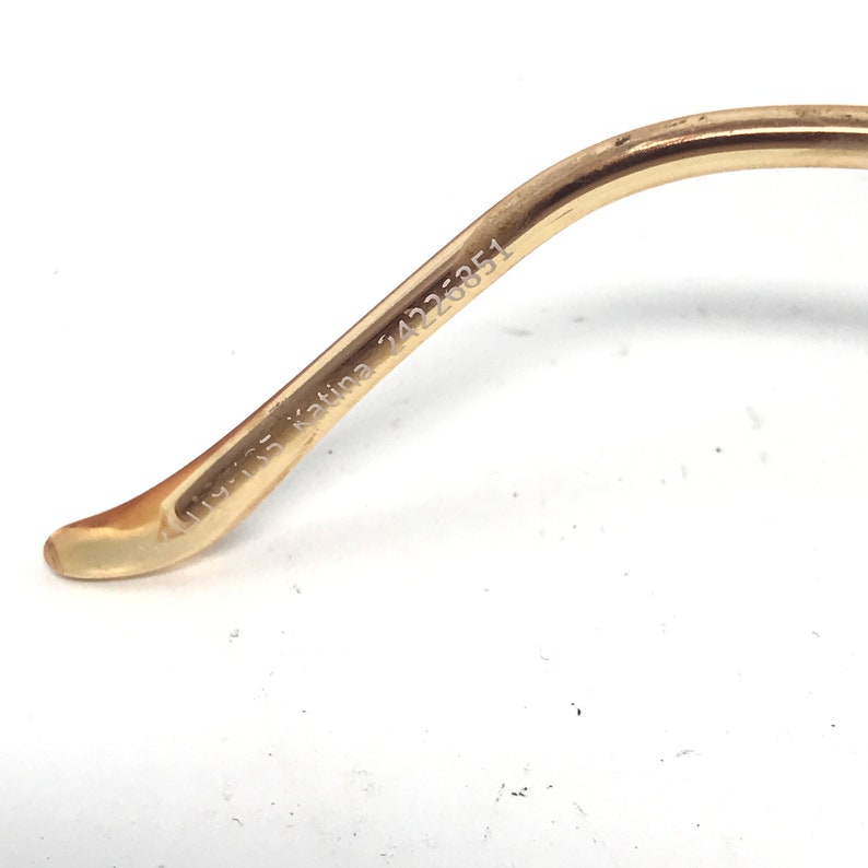 May include: Close-up of a gold-colored eyeglass frame arm. The arm has the text "119-135 Katina 24226351" printed on it. The frame is curved and has a light-colored tip.