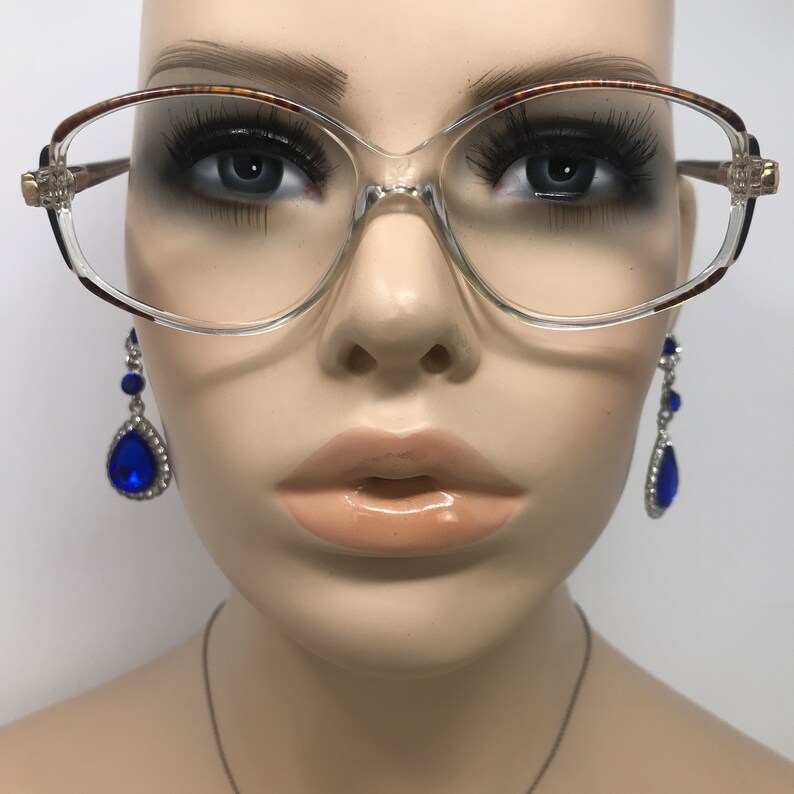 May include: Clear-framed spectacles with brown and gold detailing. The glasses have a cat-eye shape. Dangling blue teardrop earrings with silver settings are visible on either side of the face.