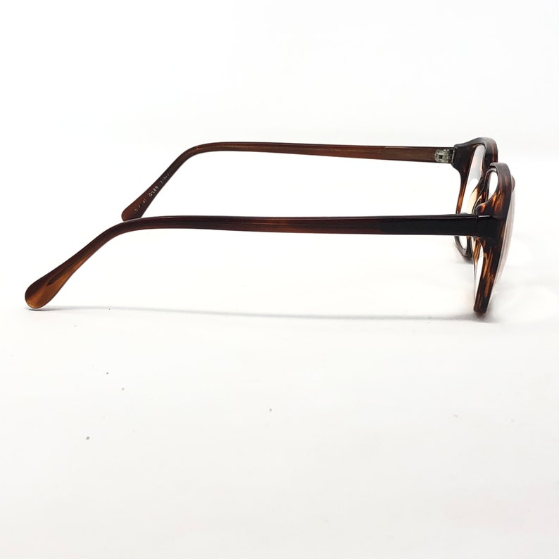 May include: A pair of eyeglasses with a brown tortoiseshell frame and temples. The glasses have a rectangular shape and are viewed from the side against a white background. The temples are thin and curve down towards the ear.