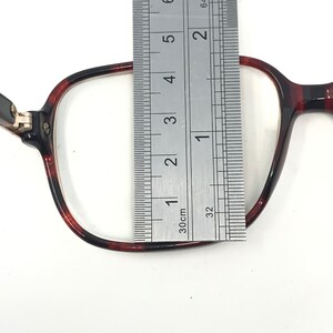 May include: Close-up of a pair of eyeglasses with a tortoiseshell pattern frame. A metal ruler is placed over the lens, measuring approximately 1.5 inches in width. The glasses are set against a white background.