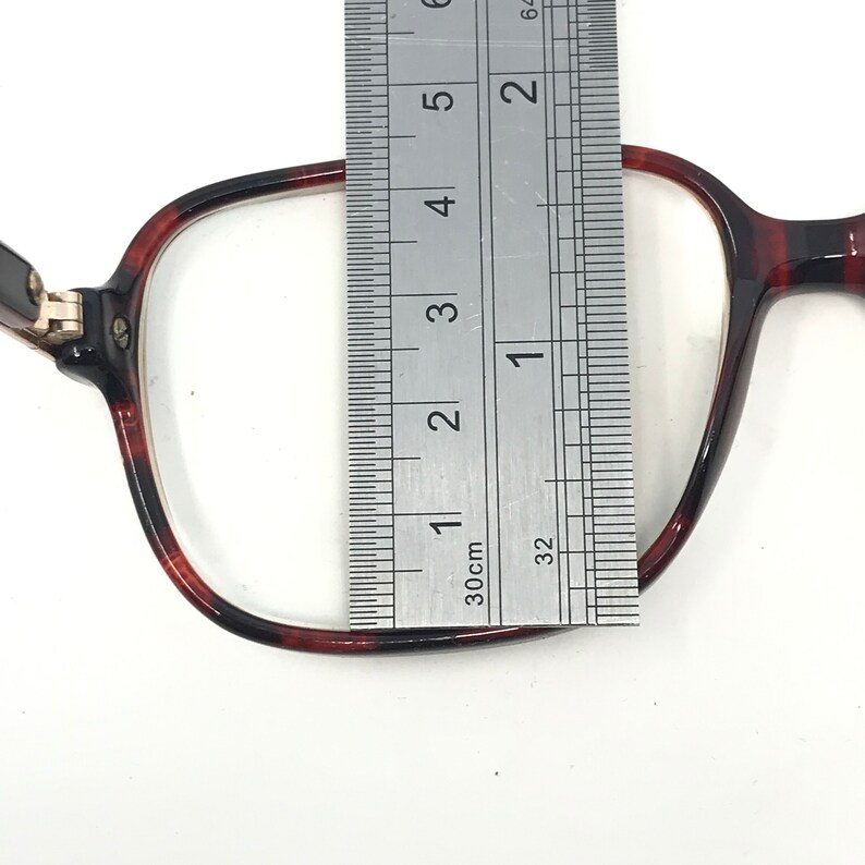 May include: Close-up of a pair of eyeglasses with a tortoiseshell pattern frame. A metal ruler is placed over the lens, measuring approximately 1.5 inches in width. The glasses are set against a white background.