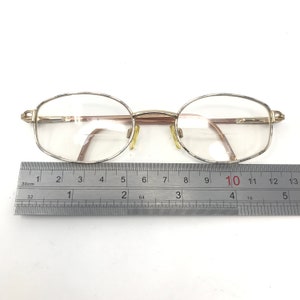 May include: Oval eyeglasses with a gold-coloured metal frame and clear lenses. The glasses are positioned above a ruler, with a measurement of approximately 13 cm across the frame. The frame has a thin, delicate design.