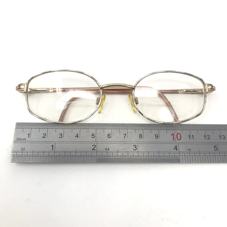 May include: Oval eyeglasses with a gold-coloured metal frame and clear lenses. The glasses are positioned above a ruler, with a measurement of approximately 13 cm across the frame. The frame has a thin, delicate design.