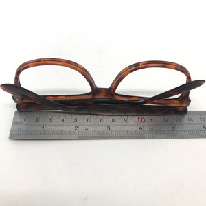 May include: Brown tortoiseshell eyeglasses with a ruler for measurement. The glasses are folded and resting on a white surface. The ruler shows measurements in inches and centimeters.