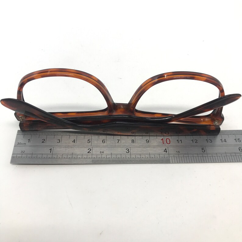 May include: Brown tortoiseshell eyeglasses with a ruler for measurement. The glasses are folded and resting on a white surface. The ruler shows measurements in inches and centimeters.