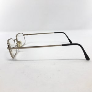 May include: Silver-framed eyeglasses with rectangular lenses. The frames have a thin, metallic appearance, with black temple arms. The glasses are positioned on a white surface, showcasing their side profile. The lenses are clear, and the nose pads are visible.