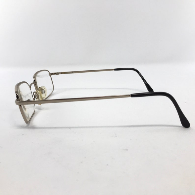 May include: Silver-framed eyeglasses with rectangular lenses. The frames have a thin, metallic appearance, with black temple arms. The glasses are positioned on a white surface, showcasing their side profile. The lenses are clear, and the nose pads are visible.