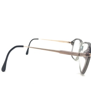 May include: A pair of spectacles with a grey and gold frame. The lenses are clear, and the arms are black with gold accents. The spectacles have a classic, angular shape.