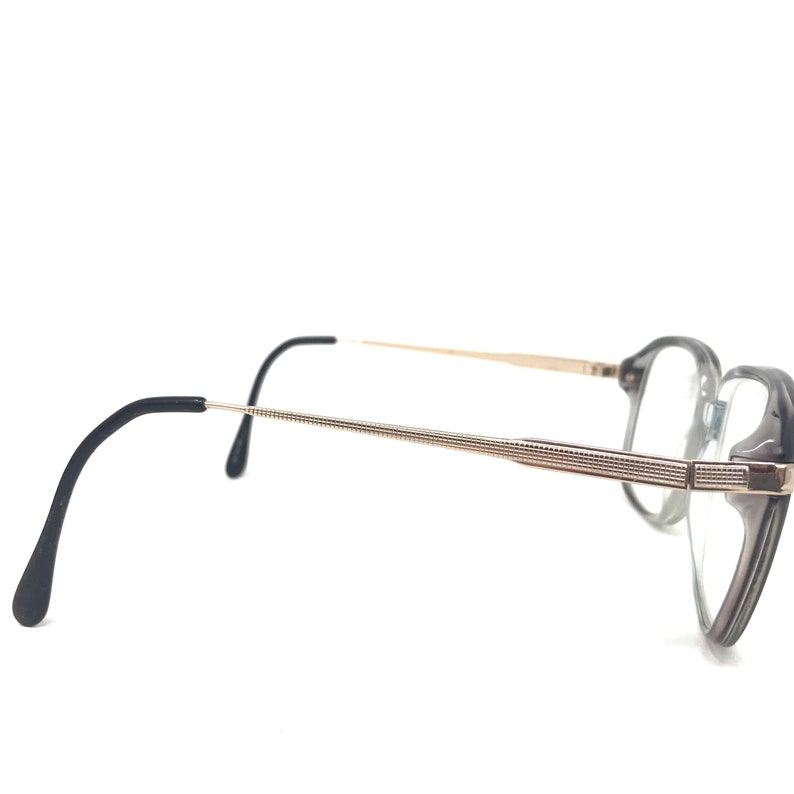 May include: A pair of spectacles with a grey and gold frame. The lenses are clear, and the arms are black with gold accents. The spectacles have a classic, angular shape.