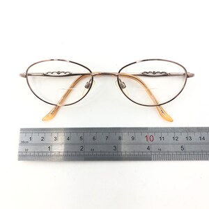 May include: Oval-shaped eyeglasses with a light brown metal frame and clear lenses. The frame features decorative detailing above the lenses. The glasses are approximately 5.5 inches wide, measured with a ruler.