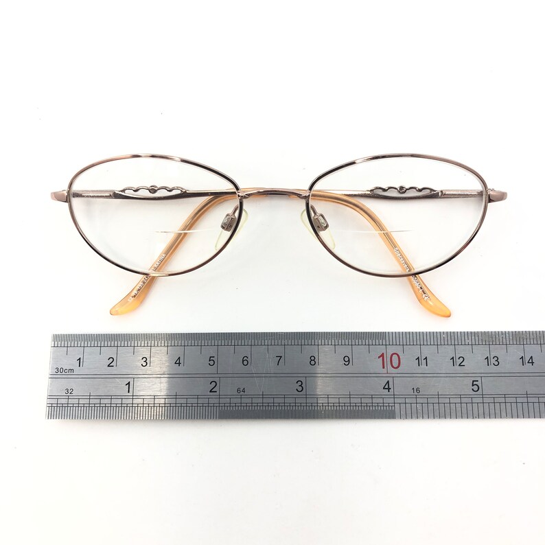 May include: Oval-shaped eyeglasses with a light brown metal frame and clear lenses. The frame features decorative detailing above the lenses. The glasses are approximately 5.5 inches wide, measured with a ruler.