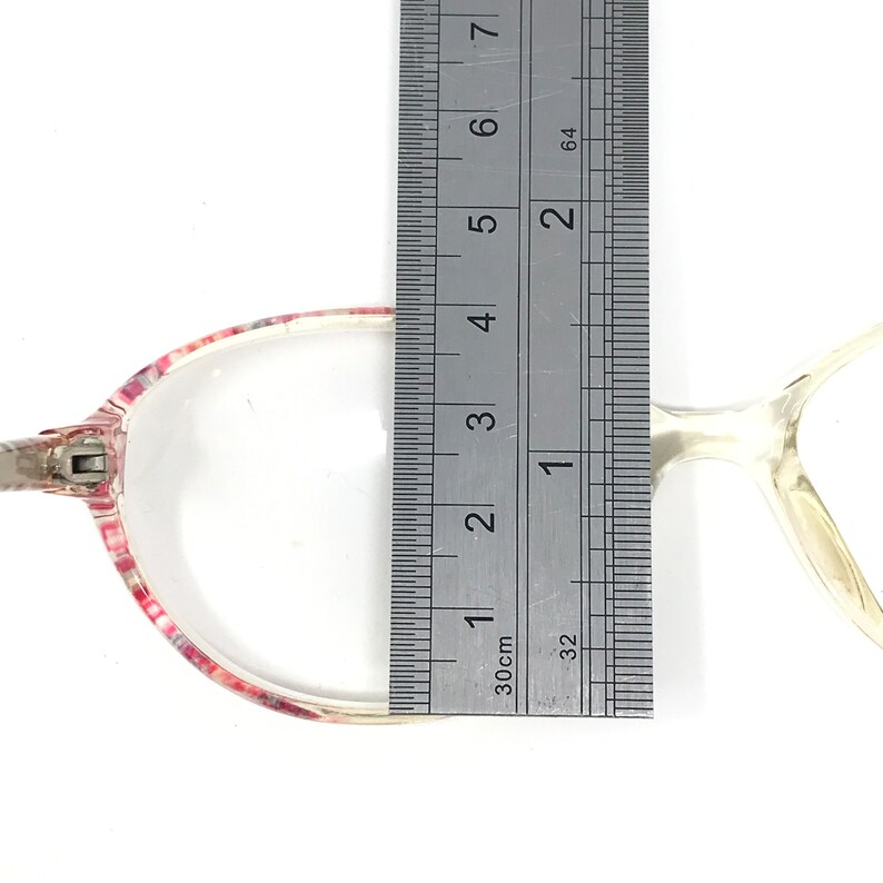 May include: Clear eyeglasses with a pink and white patterned frame. The glasses are measured with a ruler, showing a width of approximately 1.5 inches. The temples are a light, translucent color.
