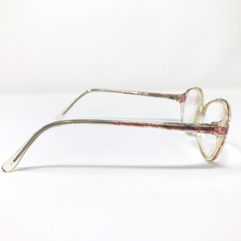 May include: Clear-framed eyeglasses with a floral pattern in pink and red. The glasses have a rounded shape and thin, silver-toned arms. The lenses are clear, and the overall design is vintage-inspired.