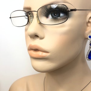 Puede incluir: Primer plano de un maniqu&iacute; con gafas rectangulares con una montura fina de color marr&oacute;n oscuro. El maniqu&iacute; tambi&eacute;n tiene pesta&ntilde;as largas y un par de pendientes colgantes azules y plateados. El fondo es blanco.