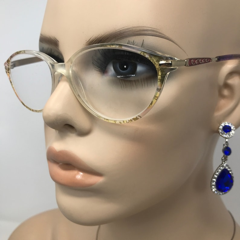 May include: Clear eyeglasses with a patterned design on the frames. The glasses are paired with dangling blue and silver earrings. The frames have a light tint and the lenses are clear.