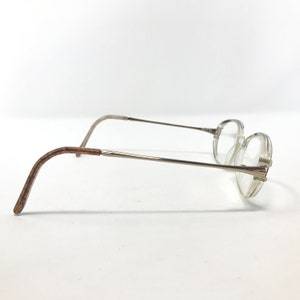 K&ouml;nnte beinhalten: Eine Brille mit klaren, runden Gl&auml;sern und d&uuml;nnen, silberfarbenen Metallrahmen. Die B&uuml;gel sind hellbraun. Die Brille ist vor einem schlichten wei&szlig;en Hintergrund platziert.