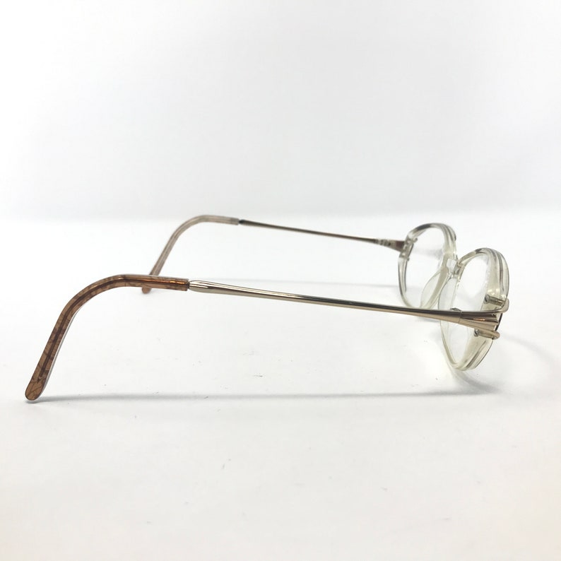 K&ouml;nnte beinhalten: Eine Brille mit klaren, runden Gl&auml;sern und d&uuml;nnen, silberfarbenen Metallrahmen. Die B&uuml;gel sind hellbraun. Die Brille ist vor einem schlichten wei&szlig;en Hintergrund platziert.