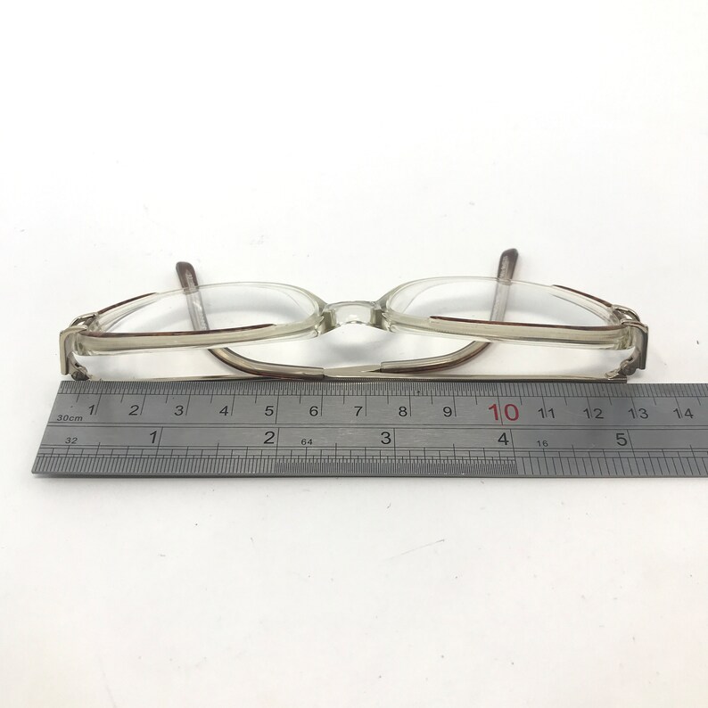 K&ouml;nnte beinhalten: Eine Brille mit transparentem Rahmen und braunen Akzenten, etwa 14 cm lang. Ein Metalllineal liegt neben der Brille auf einer wei&szlig;en Oberfl&auml;che.
