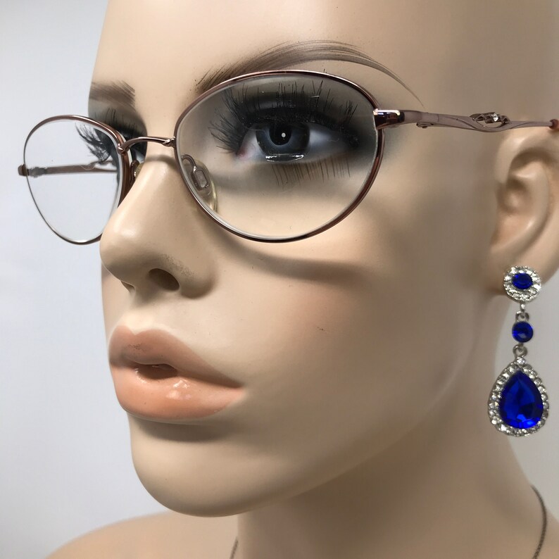 Op de afbeelding: Een paar ovale brillen met een ros&eacute;gouden montuur en heldere glazen. De bril zit op een mannequinhoofd. De mannequin draagt ​​blauwe druppelvormige oorbellen met strass.