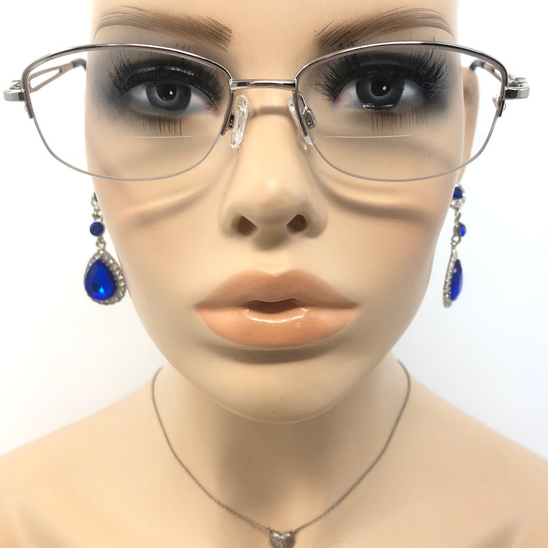 May include: A mannequin head displaying silver-framed eyeglasses and dangling blue teardrop earrings. The earrings are adorned with clear rhinestones. A silver necklace with a heart pendant is also visible.
