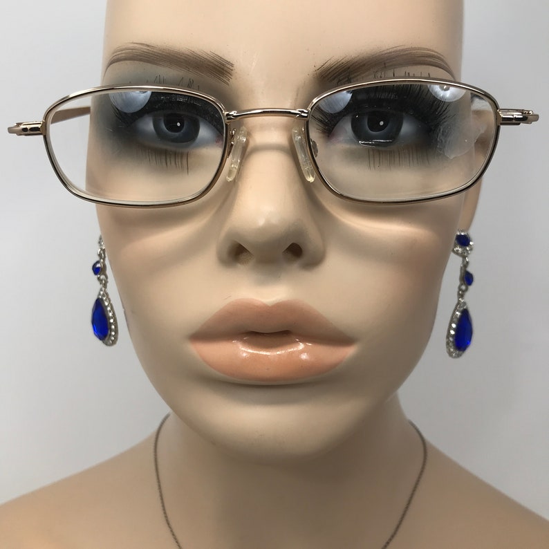 May include: Gold-rimmed eyeglasses with rectangular lenses are displayed on a mannequin. The mannequin also wears dangling earrings with blue teardrop-shaped gems and silver accents. The background is plain white.
