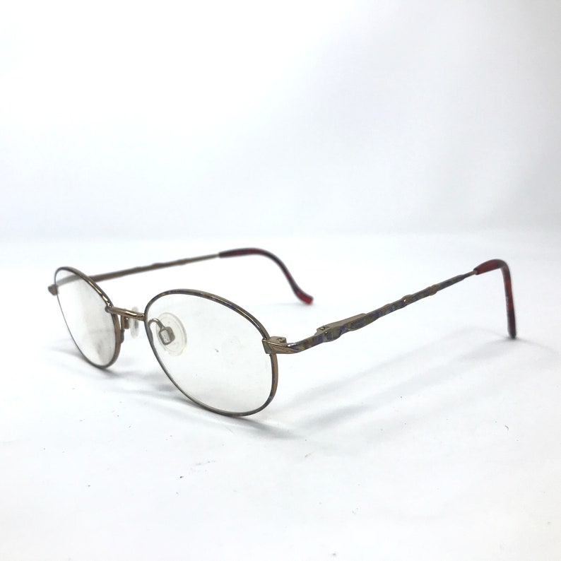 May include: A pair of gold-toned metal spectacles with oval-shaped lenses. The frames have a thin, delicate design and the arms are slightly curved.