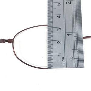 May include: Close-up of a pair of eyeglasses with a brown metal frame. The lens is clear, and a ruler is placed next to the frame, showing a measurement of approximately 1 inch.