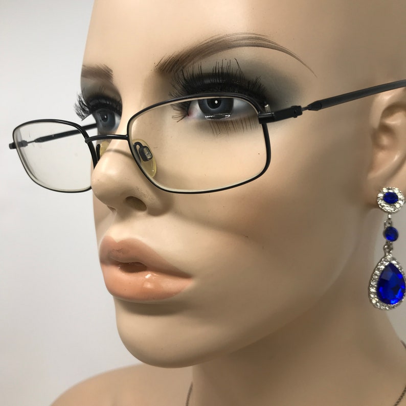 Puede incluir: Maniqu&iacute; con gafas rectangulares negras con lentes transparentes. El maniqu&iacute; tambi&eacute;n lleva pendientes colgantes con una gema azul en forma de l&aacute;grima rodeada de gemas claras m&aacute;s peque&ntilde;as. El fondo es blanco.