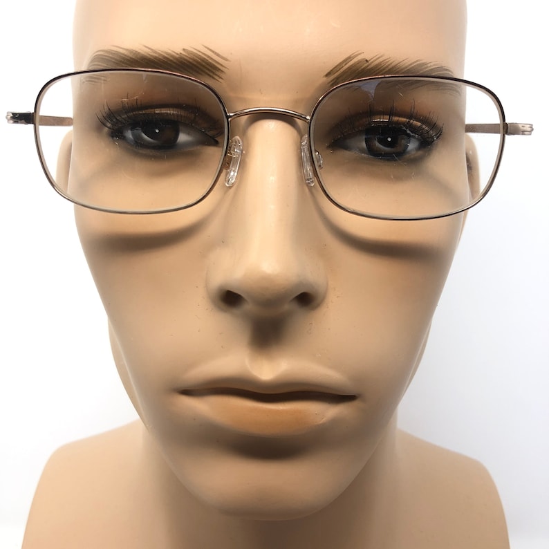 May include: Eyeglasses with a square frame made of thin, gold-coloured metal. The lenses are clear, and the nose pads are transparent. The glasses are displayed on a mannequin head with a neutral skin tone, against a white background.