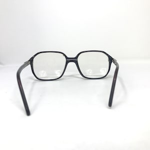May include: Black-framed spectacles with a geometric design. The lenses are clear, and the arms are a dark brown colour. The glasses are set against a white background.