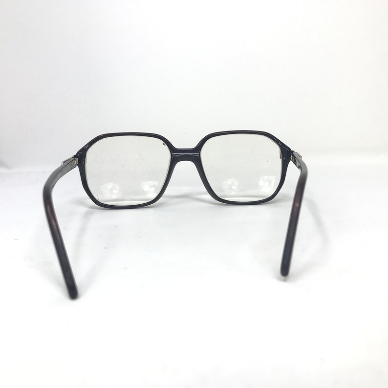 May include: Black-framed spectacles with a geometric design. The lenses are clear, and the arms are a dark brown colour. The glasses are set against a white background.