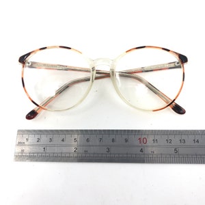 K&ouml;nnte beinhalten: Brille mit transparentem Rahmen, runden Gl&auml;sern und braunen Schildpatt-Akzenten an den oberen R&auml;ndern und B&uuml;geln. Die Brille hat einen transparenten Steg und B&uuml;gel. Der Rahmen ist etwa 14 cm breit.
