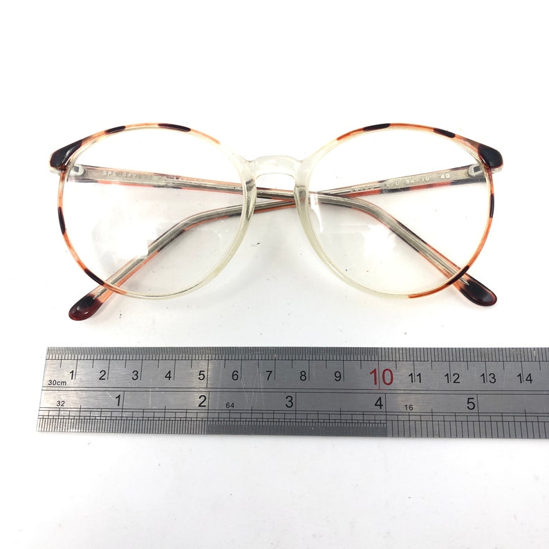 K&ouml;nnte beinhalten: Brille mit transparentem Rahmen, runden Gl&auml;sern und braunen Schildpatt-Akzenten an den oberen R&auml;ndern und B&uuml;geln. Die Brille hat einen transparenten Steg und B&uuml;gel. Der Rahmen ist etwa 14 cm breit.