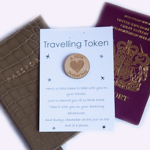 May include: A white card with the text "Travelling Token" and a small wooden token with the text "A little pocket hug" on it. The card also has a passport and a brown leather passport holder in the background.