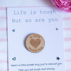 May include: A white card with the text "Life is tough But so are you" and a small wooden heart with the text "A little pocket hug" on it. The card has a small star and heart drawn on it.