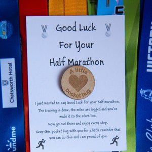 May include: A white card with black text that reads "Good Luck For Your Half Marathon". A small wooden heart with the text "A little pocket hug" is attached to the card. The card is surrounded by colourful ribbons.