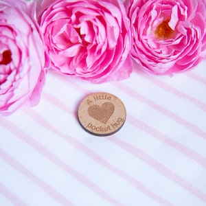 May include: A small wooden token with the text "A little pocket hug" and a heart shape engraved on it. The token is surrounded by three pink roses on a white and pink striped background.
