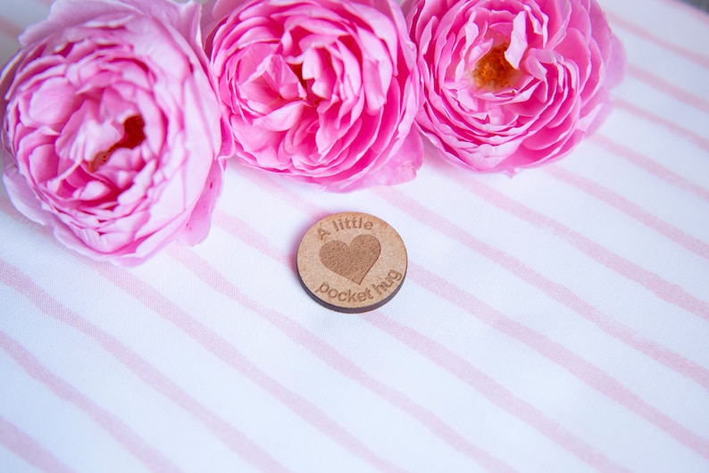 May include: A small wooden token with the text "A little pocket hug" and a heart shape engraved on it. The token is surrounded by three pink roses on a white and pink striped background.