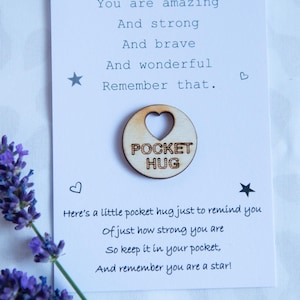 May include: A white card with a handwritten message that reads "You are amazing And strong And brave And wonderful Remember that." A small wooden circle with a heart cut out and the words "Pocket Hug" is attached to the card. The card is on a white surface with lavender flowers.