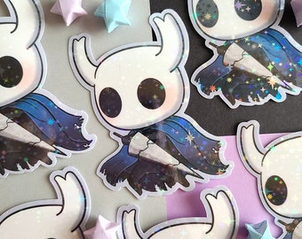 May include: A collection of stickers featuring a cartoon character with a white head, large black eyes, and a blue cape. The stickers have a holographic, sparkly finish. Origami stars in pastel colors are scattered around the stickers.