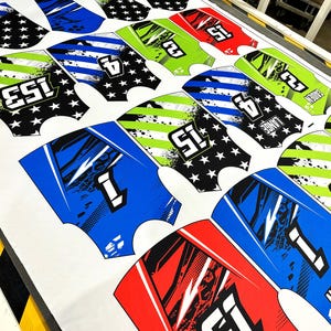 K&ouml;nnte beinhalten: Bedruckte Stoffst&uuml;cke mit verschiedenen Designs, darunter Sterne, Streifen und Zahlen. Die Farbpalette umfasst Blau, Gr&uuml;n, Rot und Schwarz. Der Stoff ist wahrscheinlich f&uuml;r Sportbekleidung oder Sportuniformen bestimmt.